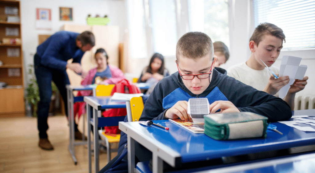 Students working independently at their desks while teacher helps in the background.