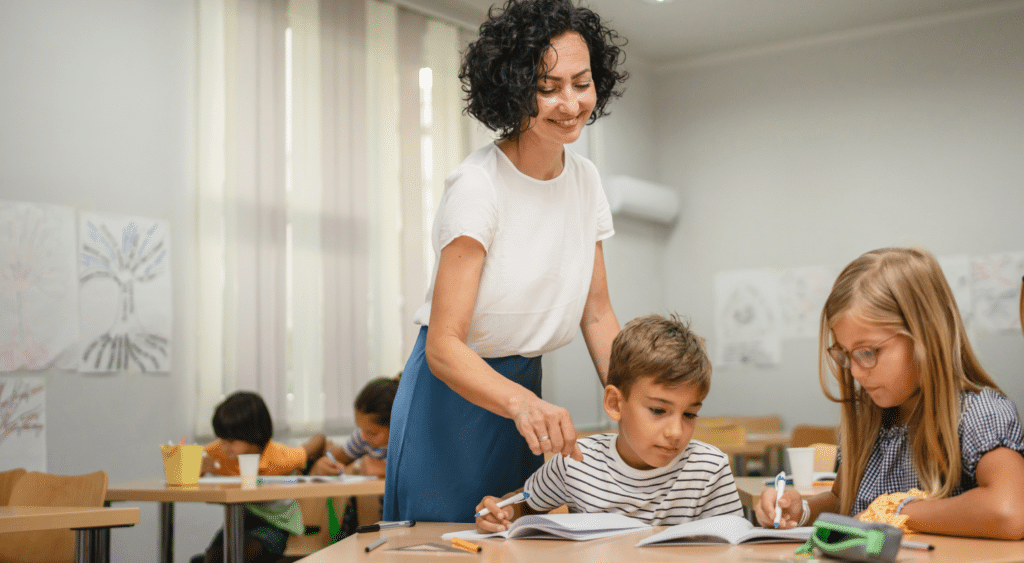 Two students working at a table with substitute teacher standing by helping