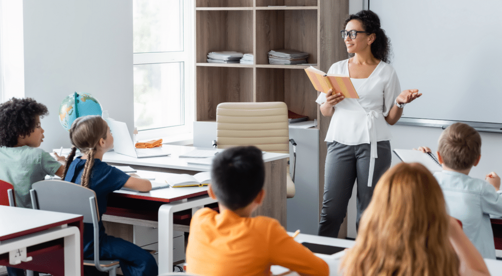 Teacher reading a book at the front of the classroom as students watch and listen.