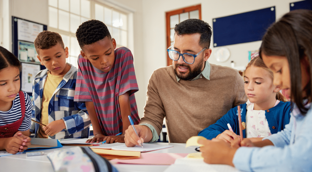 Teacher writing at a desk with students gathered around watching